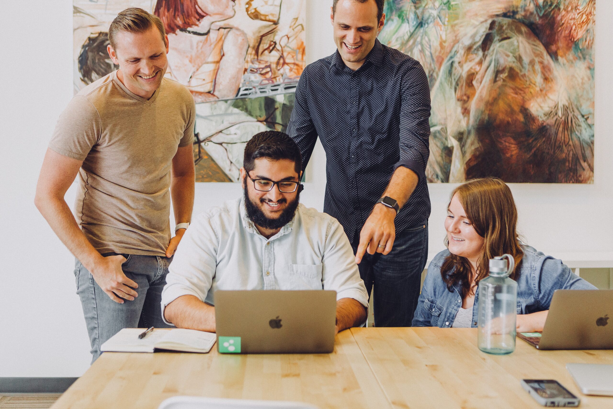 4 people are gathered around a computer smiling, two sat at a large desk, the other two stood behind, one of them pointing to something on the screen. Image represents employee recognition. Image by Jud Mackrill via Unsplash.