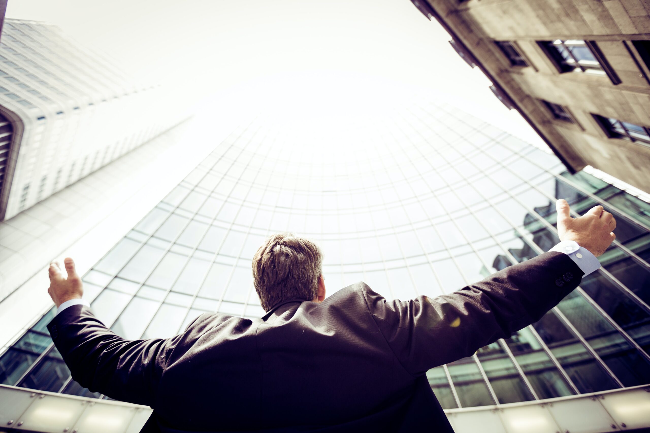 Man wearing a suit with greyed hair opens his arms up towards a tall skyscraper, the top of which is obscured by the blazing sun, representing his future career development opportunities. Photo by Razvan Chisu via Unsplash.