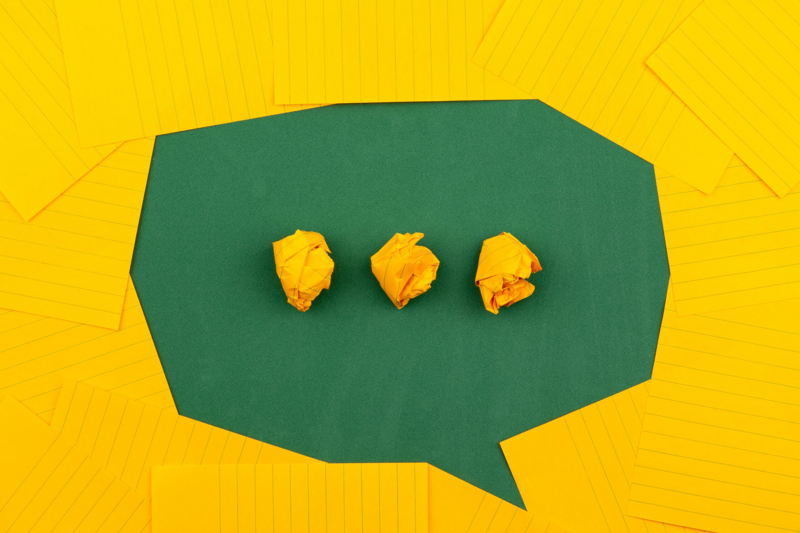 Picture of a speech bubble created using negative space – there is a green background carved into the shape of a bubble by a lot of yellow lined pieces of card. Inside the speech bubble, there are three scrumpled pieces of this yellow card which form an ellipsis. Photo by Volodymyr Hryshchenko on Unsplash