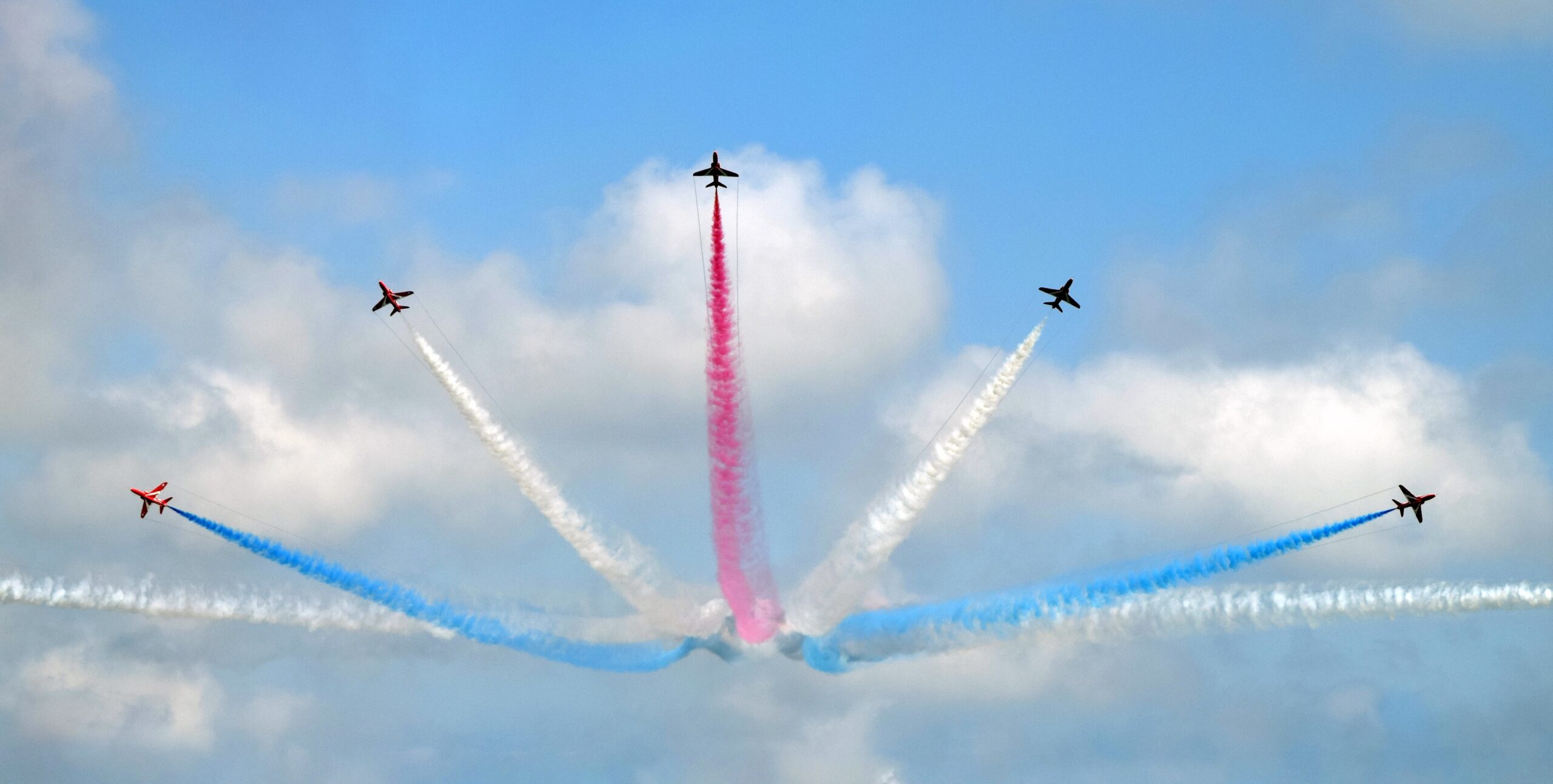 5 Red Arrow jets flying in a formation where they fan out from a singular point, trailing red, white and blue smoke behind them. This image represents the power of a strong company culture in creating synchronicity and a sense of unity. Image by Owen Kemp via Unplash: https://unsplash.com/photos/I8BaNbAd9os?utm_source=unsplash&utm_medium=referral&utm_content=creditShareLink